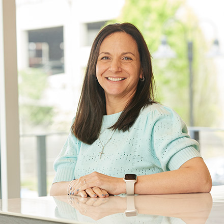 Woman with straight dark brown hair, wearing a light blue patterned blouse and a smartwatch, seated at a table with hands folded, smiling warmly in a bright indoor environment.