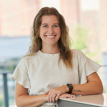 Young woman with wavy light brown hair, wearing a white short-sleeve top and an Apple Watch, smiling broadly while resting her arms on a surface in a softly lit indoor space.