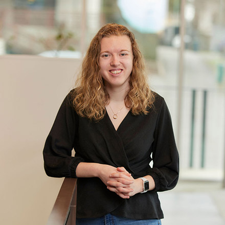 Young woman with curly light brown hair, wearing a black long-sleeve top and an Apple Watch, standing and leaning slightly on a railing with hands clasped, smiling gently in an indoor setting.