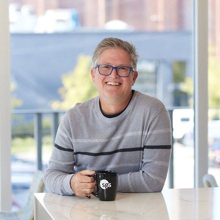 Middle-aged man with short gray hair and glasses, wearing a gray striped sweater, seated at a table holding a black coffee mug, smiling warmly in a well-lit indoor space.