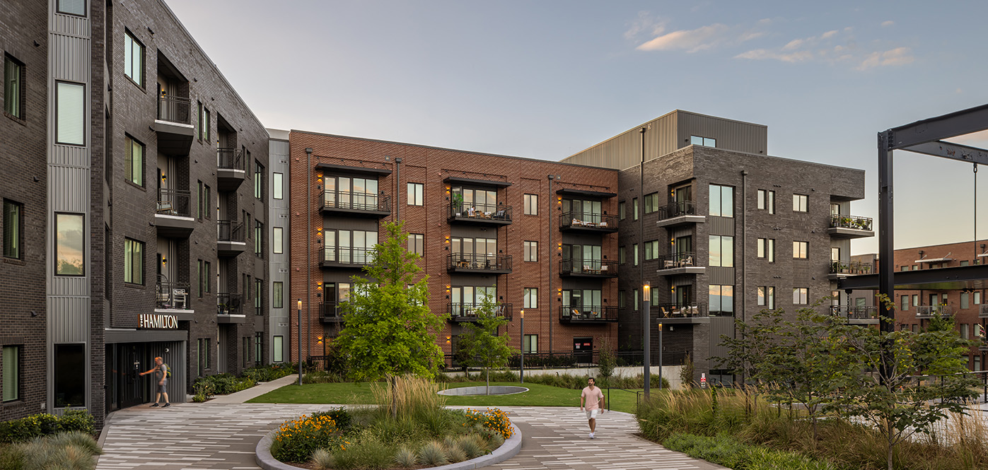 Serene apartment courtyard at sunset with red and gray brick façades, balconies, trees, and landscaped walkways in front of The Hamilton apartments.