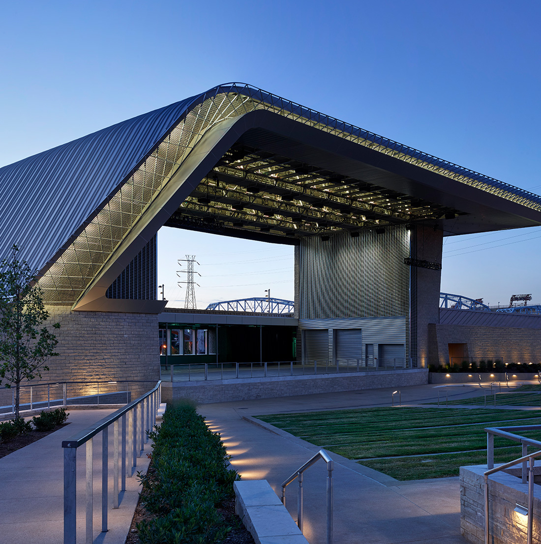 A modern amphitheater with dramatic roof architecture is shown empty at dusk, with pathways, greenery, and accent lighting.