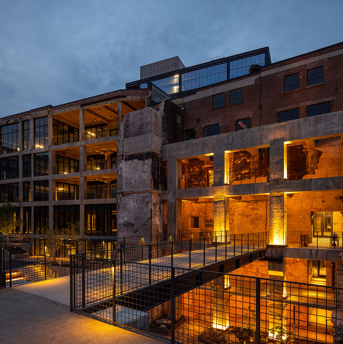 Exterior view of a modern architectural renovation combining exposed brick and concrete with glass walls, illuminated warmly at dusk, highlighting the blend of historic and contemporary design elements with a metal walkway and safety railing in the foreground.