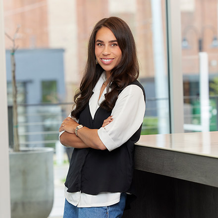 Young woman with long dark brown hair wearing a white blouse under a black vest, standing by a counter with arms crossed, smiling confidently.