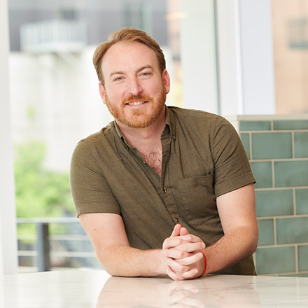 A man with short red hair and a beard sits at a light-colored countertop, leaning forward with his hands clasped. He is wearing a short-sleeved olive-green shirt and smiling at the camera. Behind him is a large window with a view of an urban setting and a tiled wall to the right.