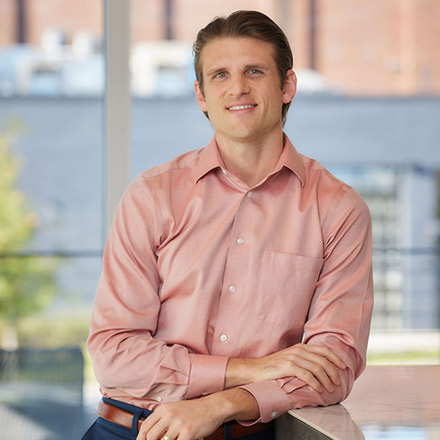 Man with short light brown hair wearing a salmon pink button-down shirt, leaning on a table with arms crossed, smiling in a bright indoor setting.