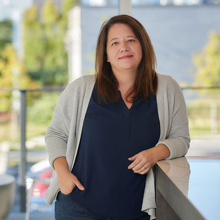 Woman with shoulder-length dark brown hair, wearing a navy blue top and light gray cardigan, standing with one hand in pocket and the other resting on a table, smiling gently.