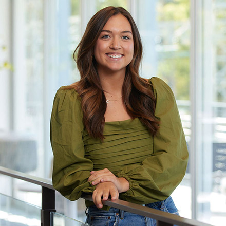 Young woman with long wavy brown hair, wearing an olive green blouse with puffed sleeves, leaning on a railing and smiling warmly in a well-lit indoor space.