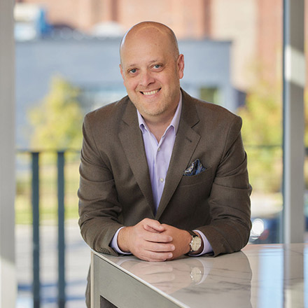 Bald man wearing a brown suit jacket and light purple shirt, seated with hands folded on a table, smiling confidently.