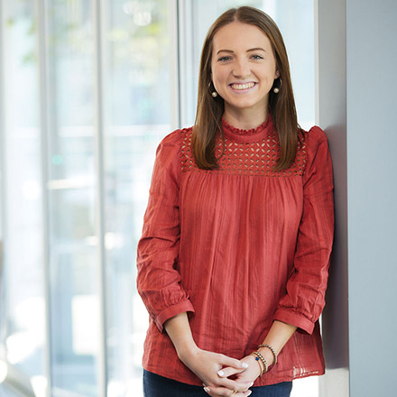Young woman with long brown hair, wearing a red blouse with lace details, standing and smiling brightly in a sunlit indoor area.