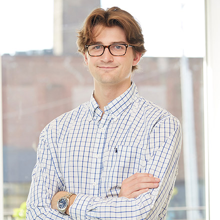 Young man with light brown hair and glasses wearing a white checkered button-down shirt, standing with arms crossed, smiling in a well-lit indoor space.