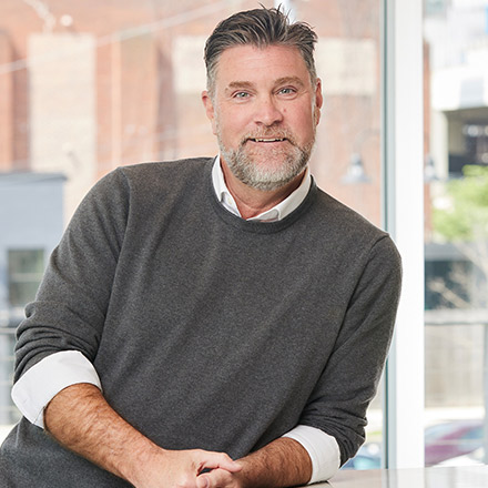 Middle-aged man with short salt-and-pepper hair and beard, wearing a dark gray sweater over a white collared shirt, sitting and smiling at a table near a window.