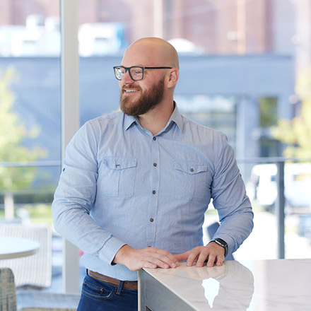 Bald man with a beard wearing glasses and a light blue button-down shirt, standing with hands resting on a countertop, looking to the side with a smile.