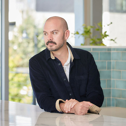 Bald man with mustache and goatee, wearing a dark button-up shirt with tan collar, seated at a table with hands clasped, looking pensively to the side.