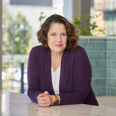Woman with shoulder-length auburn hair wearing a purple cardigan over a white top, seated at a table with hands clasped, looking directly at the camera with a neutral expression.