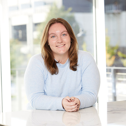 Young woman with shoulder-length brown hair wearing a light blue sweater, seated at a white table near a window, smiling gently at the camera.
