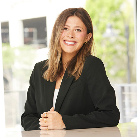 Young woman with light brown hair wearing a black blazer and white top, seated at a table, smiling brightly at the camera in a well-lit room.