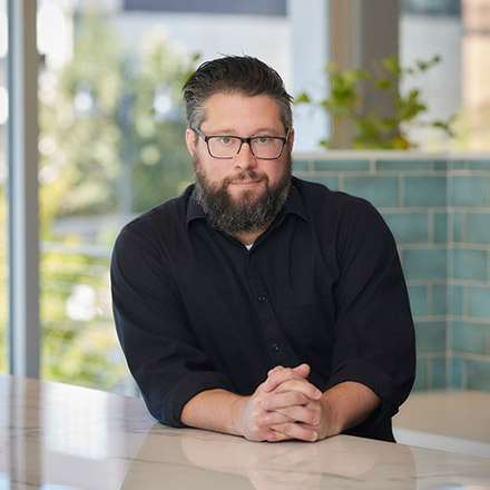 Man with short dark hair, beard, and glasses wearing a black shirt, seated at a table with hands clasped, looking attentively at the camera.
