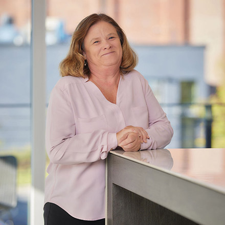 Middle-aged woman with shoulder-length light brown hair wearing a pale pink blouse, standing and leaning against a table, smiling softly.