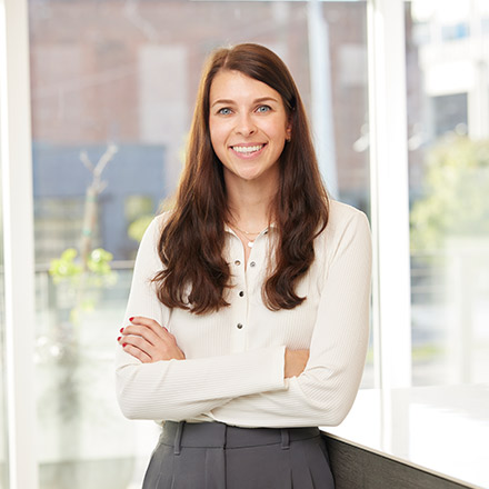 Young woman with long dark brown hair wearing a white blouse, standing near a table with arms crossed, smiling at the camera.
