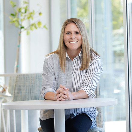 Woman with shoulder-length blonde hair wearing a striped button-down shirt, seated at a round white table, smiling at the camera.