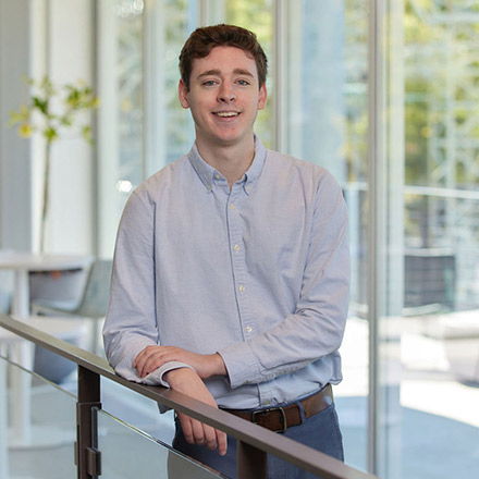 Young man with short curly red hair wearing a light blue button-down shirt, standing by a railing, smiling at the camera.