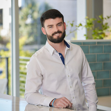 Young man with short dark hair and beard, wearing a white dress shirt, seated near a window, smiling gently.