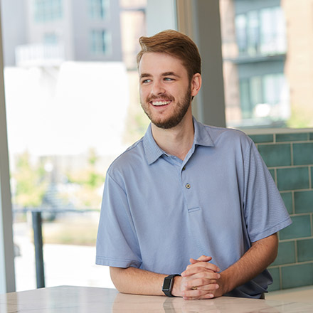 Young man with light brown hair and beard, wearing a light blue polo shirt, seated at a white table, looking to the side and smiling.
