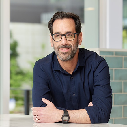Man with dark hair, beard, and glasses, wearing a navy blue shirt, seated at a white table near a window, smiling softly.