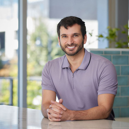 Man with short dark hair and beard, wearing a light purple polo shirt, seated near a window, smiling at the camera.