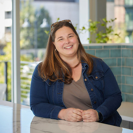 Woman with long brown hair wearing a dark blue denim jacket, seated at a white table near a window, smiling broadly.