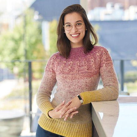Woman with straight shoulder-length brown hair and glasses wearing a multi-colored knit sweater, smiling and leaning on a counter, indoors with large windows behind.
