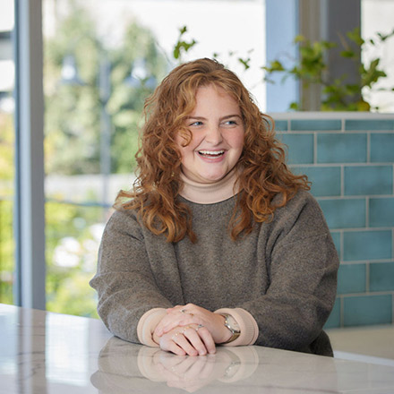 Woman with curly red hair wearing a brown sweater, laughing and seated with arms crossed on a counter, indoors with windows behind.