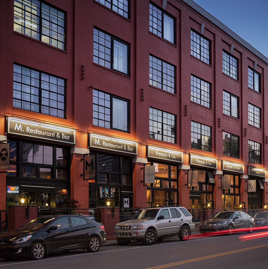 Wider evening view of the red brick building with lit signage for various restaurants and shops, multiple parked cars, and a few people visible near the entrances.