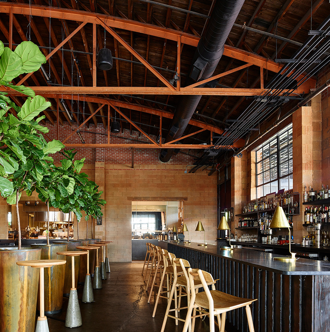 Interior view of a trendy industrial bar with exposed wood beam ceiling, brick and cinderblock walls, sleek bar seating, and large potted plants adding greenery to the space.