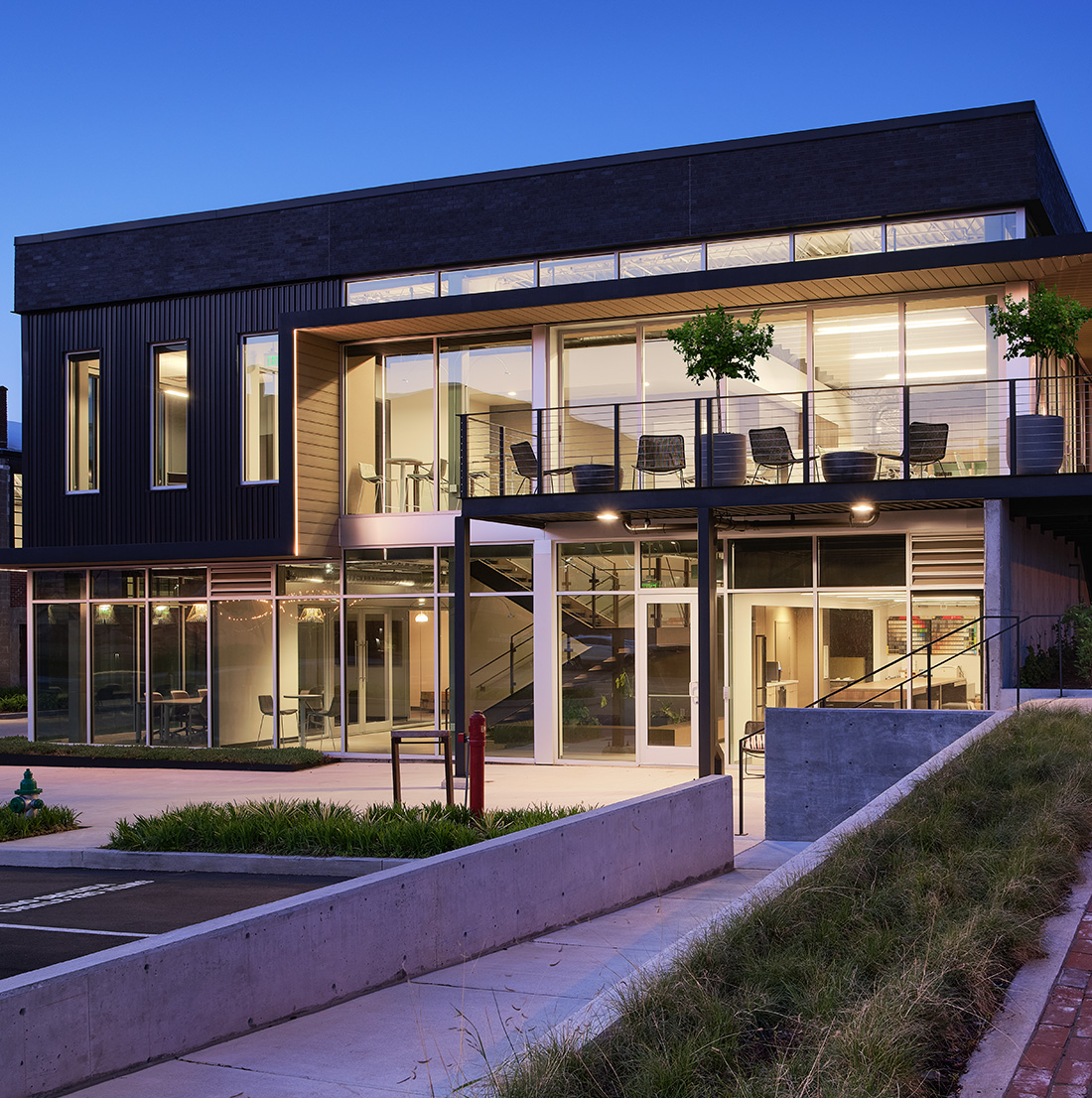 A contemporary glass and steel building is illuminated at twilight, with upper-level balconies, interior lighting, and clean landscaping enhancing its sleek design.