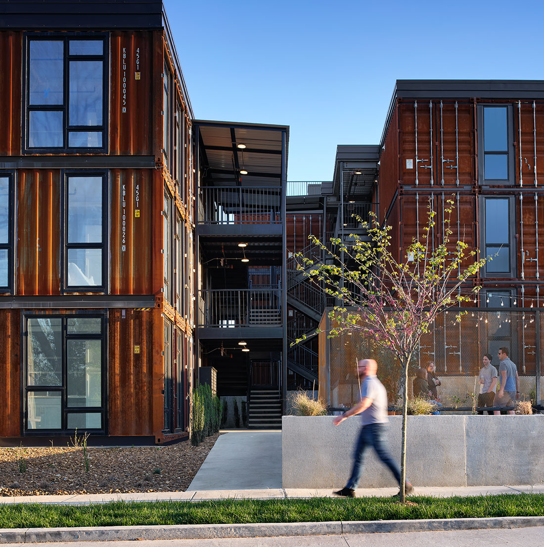 Street-level view of the residential complex exterior during daytime, showing three stories of container modules with black-framed windows and outdoor staircases. A pedestrian walks along a sidewalk in front of the building.