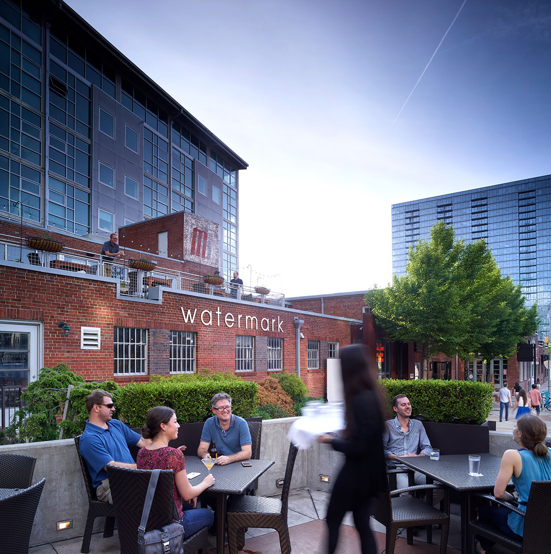 Outdoor dining area at Watermark restaurant with guests seated at patio tables beside a red brick building. A modern residential tower rises in the background, and a server moves through the scene.
