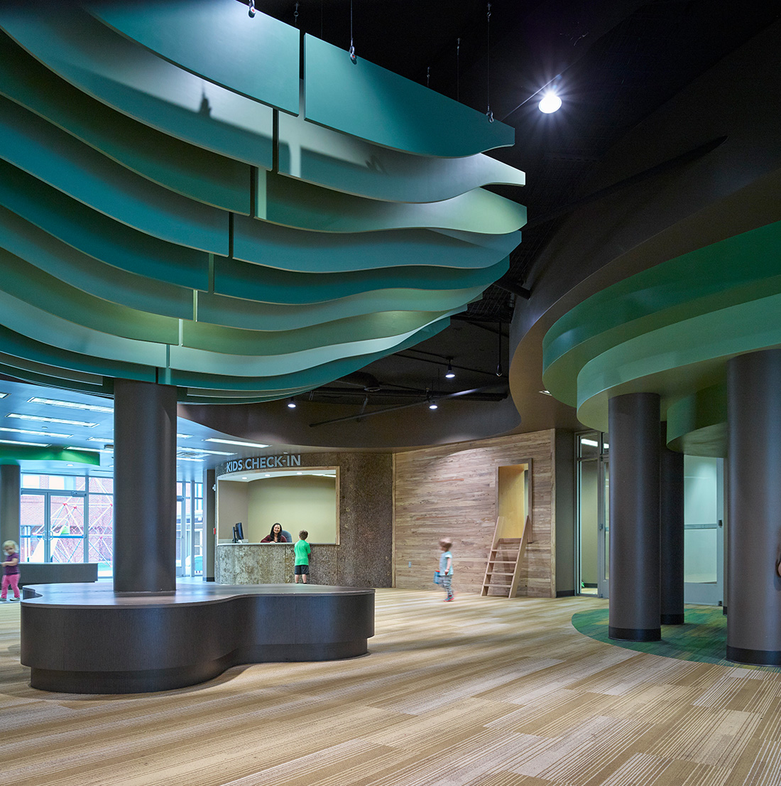 A modern children's check-in area with layered green ceiling panels, wood accents, and kids interacting near a staffed desk.