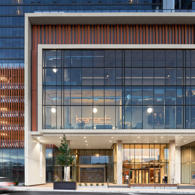 Centered front view of the building’s glass entrance with visible lobby and interior signage that reads “Kenect.” Two people are walking near the entrance.