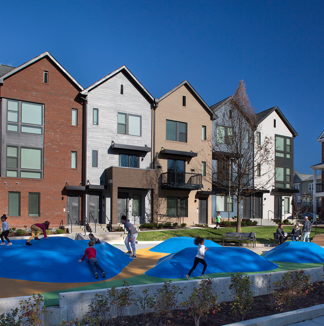 Colorful, modern townhomes with a combination of brick and siding facades face a landscaped outdoor play area. Children are shown climbing and running on soft, blue rubber mounds. The space includes benches and trees, creating a vibrant, family-friendly environment.