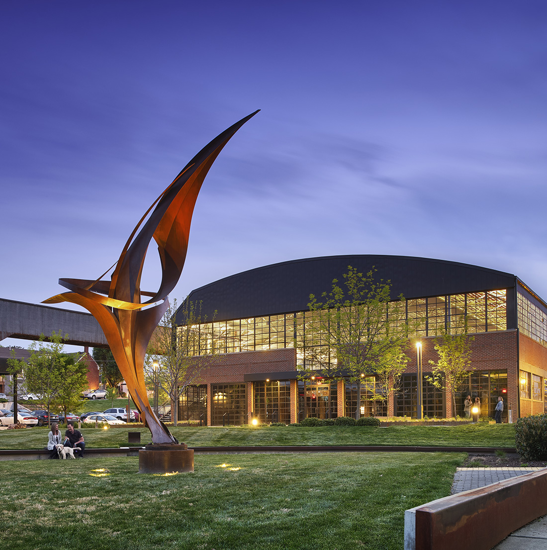 Twilight view of an abstract metal sculpture with curved forms installed on a grassy plaza. In the background, an industrial-style building with tall glass windows is illuminated from within. The surrounding area includes trees, walking paths, and people enjoying the public space.