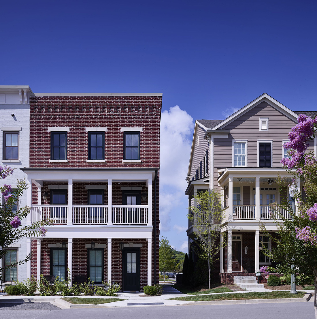 Side-by-side view of two residential homes—one red brick townhouse with double front porches and one taupe-colored traditional house with white trim and flowering trees.