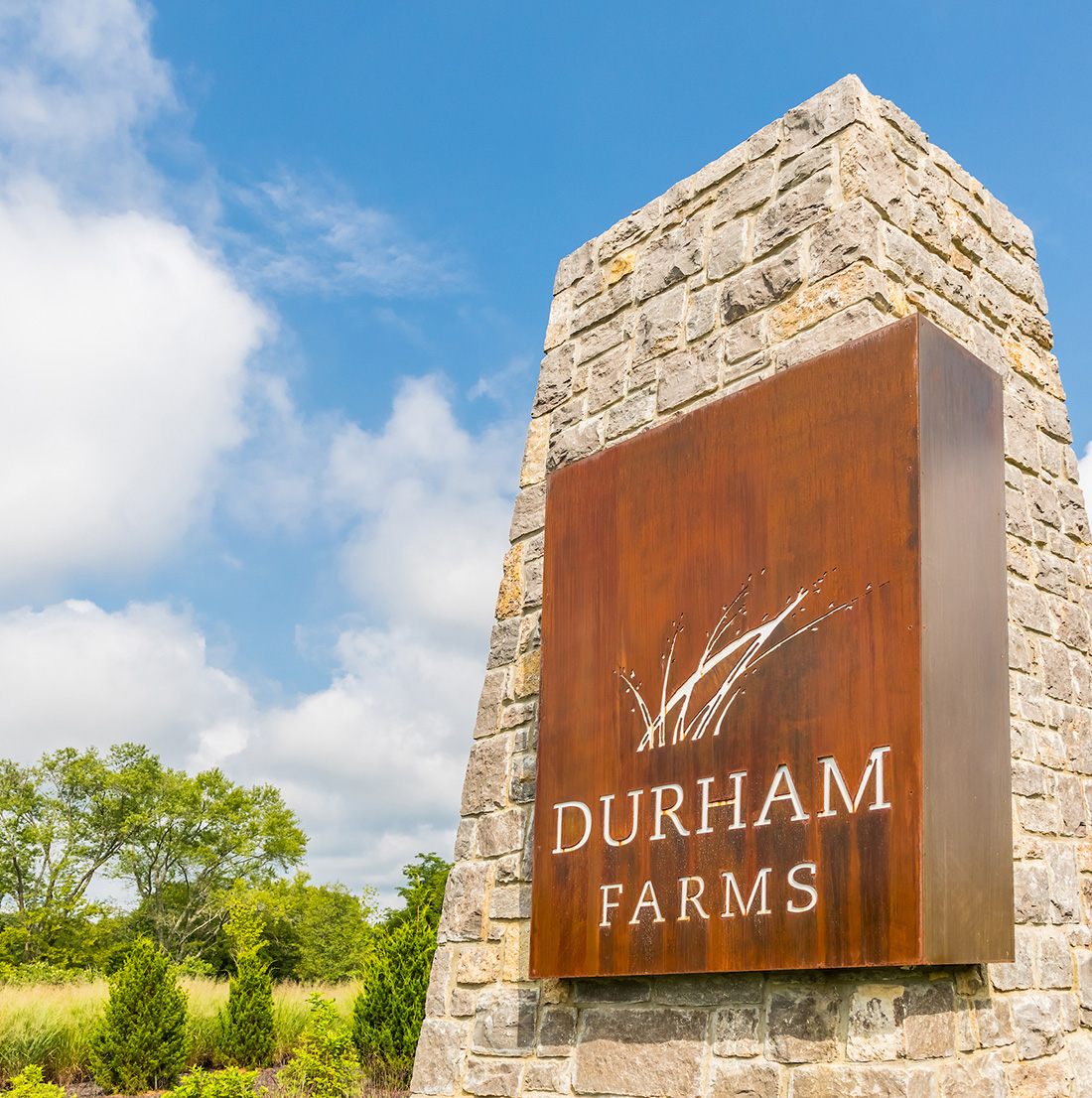 Close-up of the Durham Farms entrance sign made from stacked stone and weathered steel, set against a blue sky and surrounding greenery.
