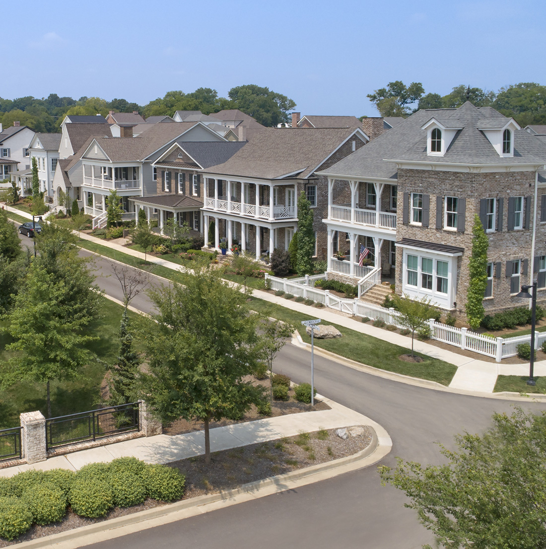 A picturesque suburban neighborhood features a row of large, elegant houses with front porches and manicured lawns lining a quiet, curved street. On the opposite side, a lush green park with trees, walking paths, and open grassy areas creates a serene, community-friendly atmosphere. Rolling hills and dense forest can be seen in the distance under a clear blue sky.