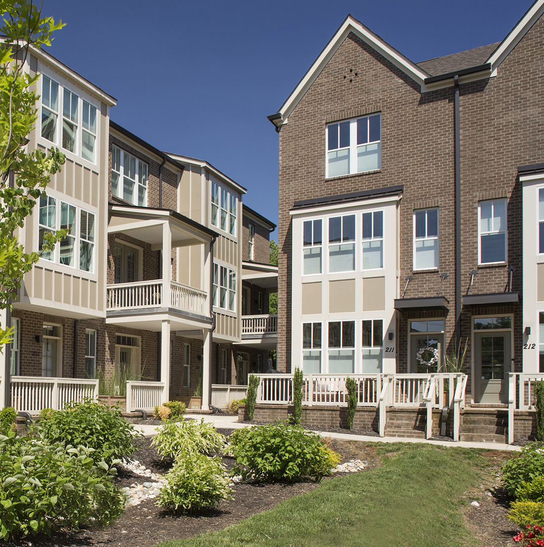 View of a landscaped courtyard surrounded by three-story townhomes with brick facades, large windows, and white trim. A couple walks along the central pathway beneath sunny skies.
