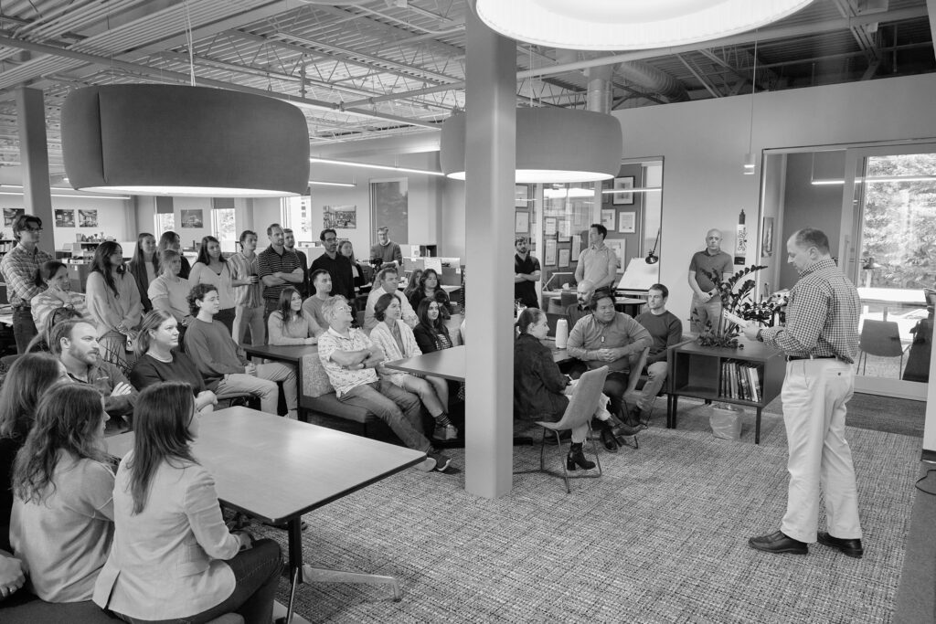 Black and white photo showing a group of people attentively listening to a speaker in a modern office setting, with several seated and standing individuals visible.