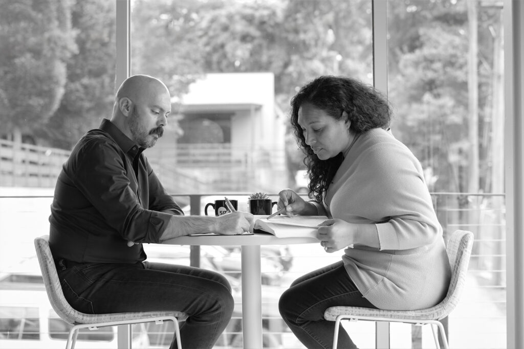 Black and white image of a man and woman seated at a small table engaged in focused discussion, with large windows and outdoor greenery visible in the background.