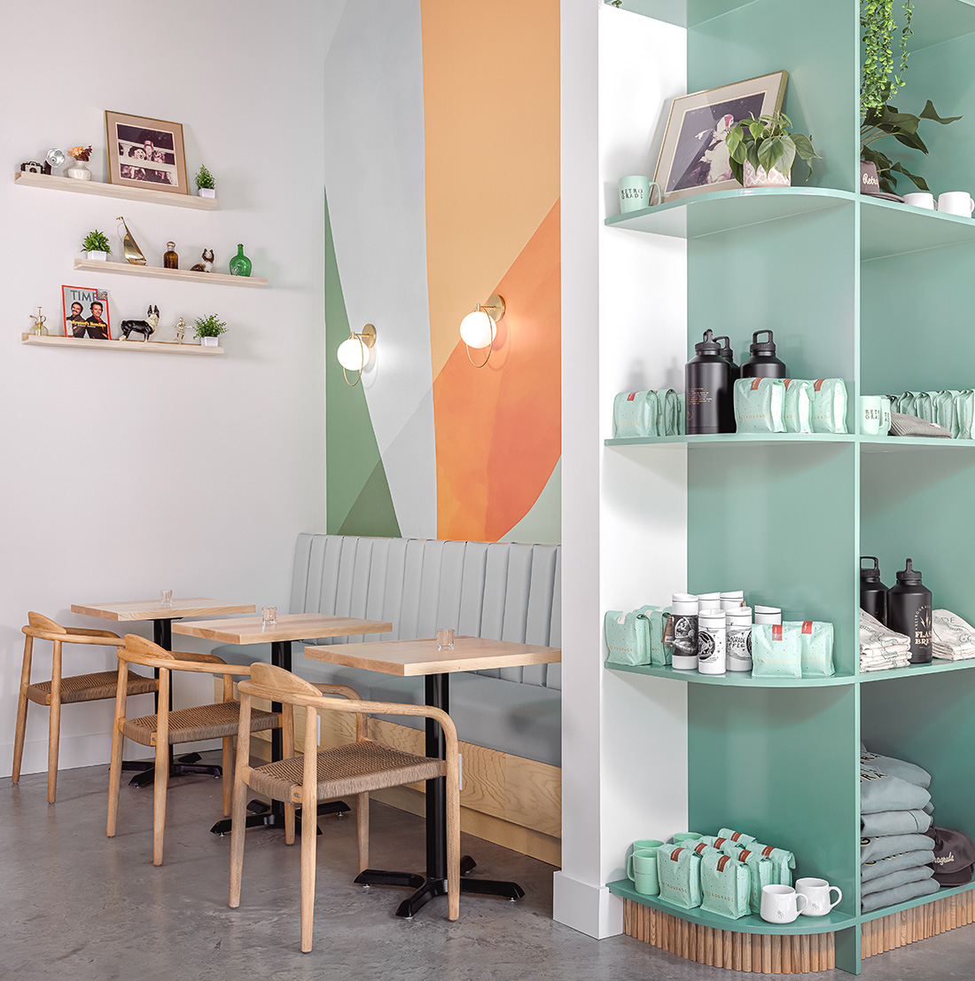 Cozy café nook with light wood tables and chairs, mint green banquette seating, a geometric mural in soft pastels, and display shelves featuring coffee merchandise and decorative items.