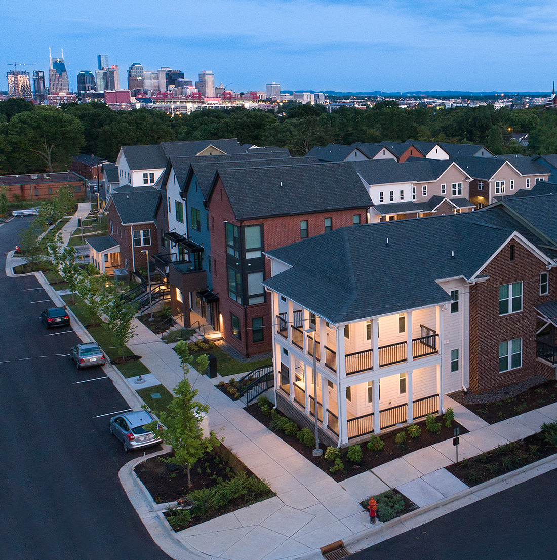 A modern suburban neighborhood features rows of townhomes and single-family houses with front porches, clean sidewalks, and neatly landscaped yards. The homes are a mix of brick and siding, illuminated warmly as evening falls. In the distance, the downtown skyline rises above the treetops, adding an urban backdrop to this peaceful residential setting.